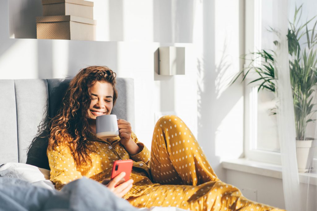 woman in yellow pajama sitting on bed and drinking coffee and looking to the phone at the sunny morning bedroom