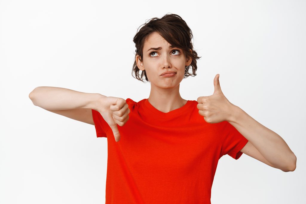 indecisive girl showing thumbs up and thumbs down looking puzzled cannot make choice weighing pros and cons white background